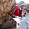 Two students take notes on clip boards while a third uses a ruler to measure a section of rock during a geology field camp.