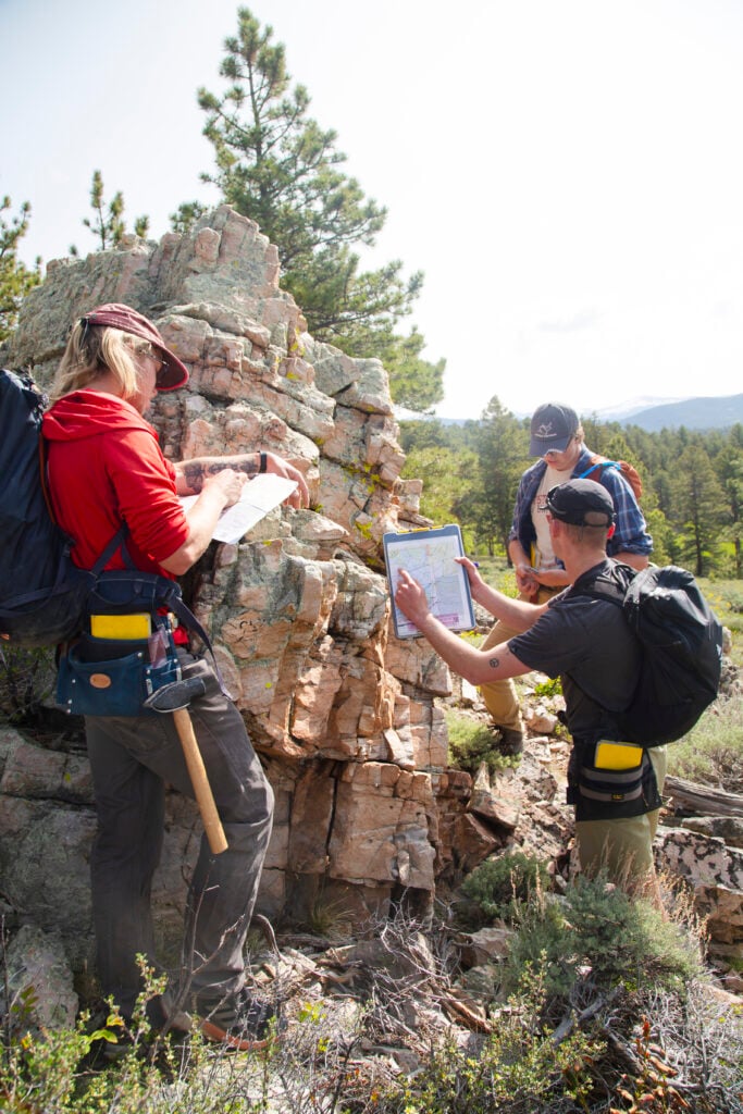 Two students examine a face of rock while a third takes notes during a geology field camp.