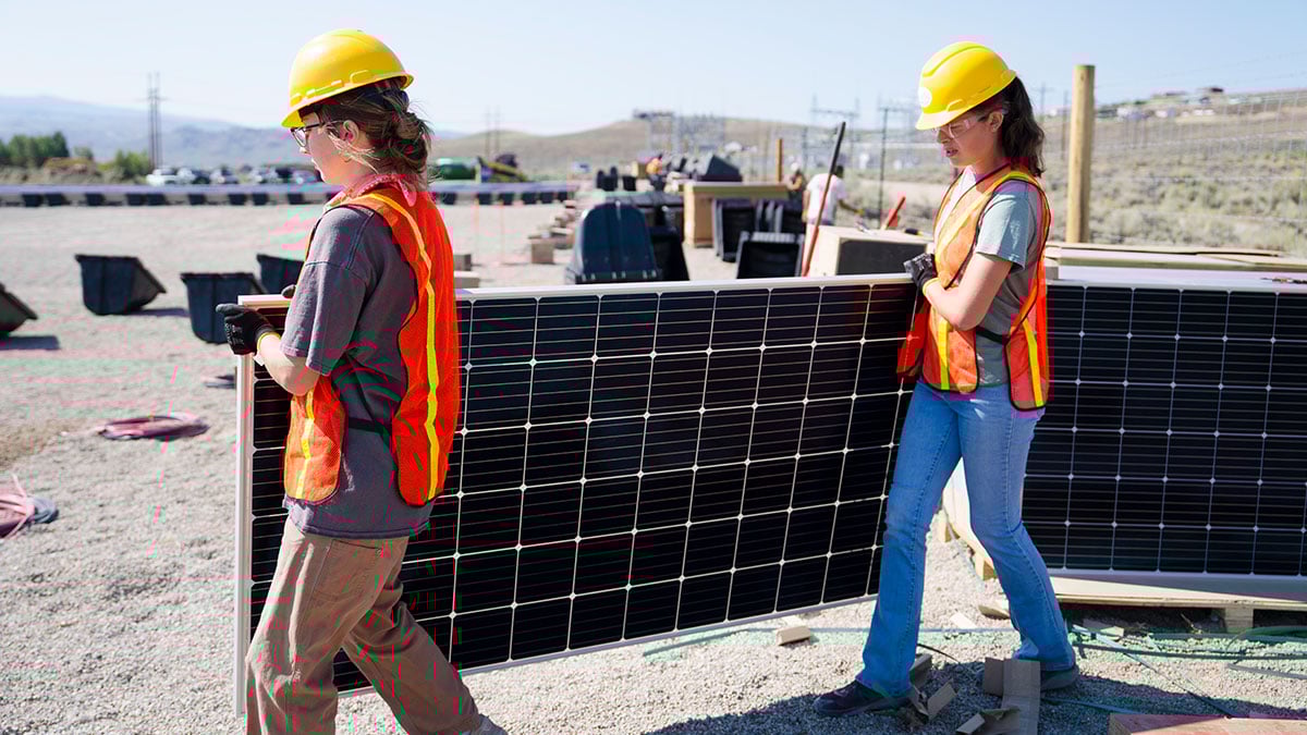 Two people carry a solar panel while helping build a solar array for Powerfield Energy.