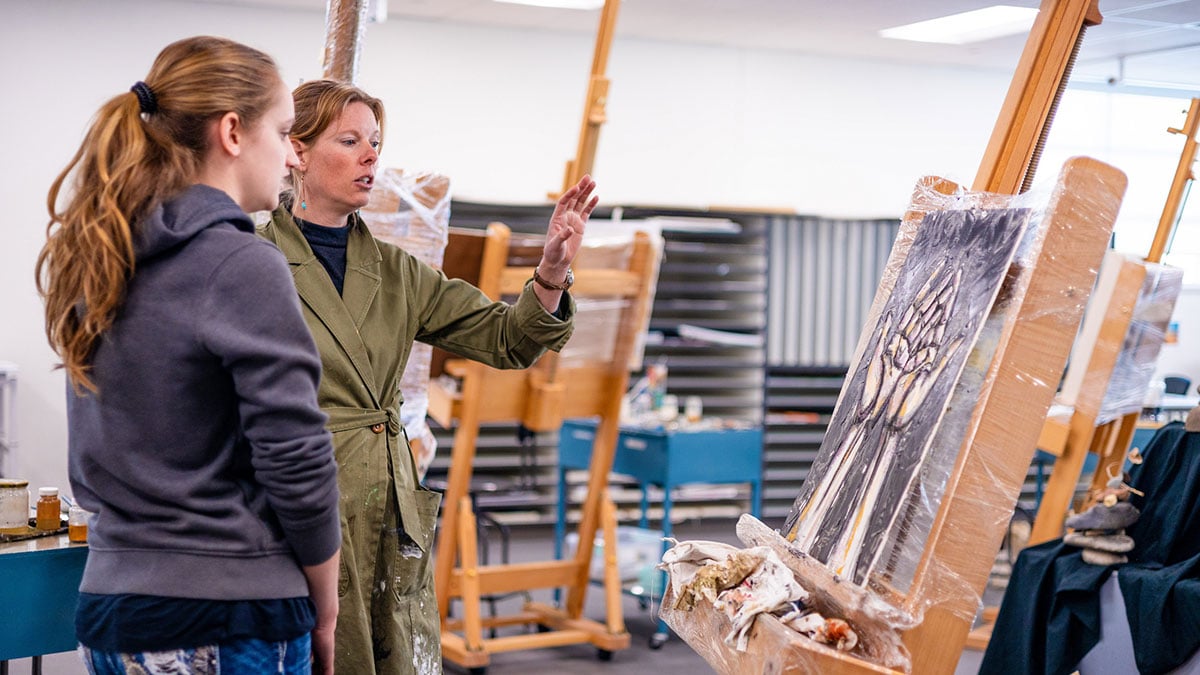 A professor gives feedback to her student next to her on her painting