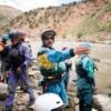 A group of river rescuers in safety gear stand on rocky riverbank, with one person gesturing toward the water as they prepare for a rescue drill.
