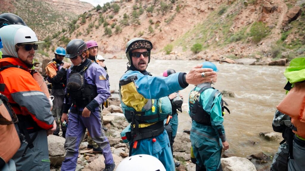 A group of river rescuers in safety gear stand on rocky riverbank, with one person gesturing toward the water as they prepare for a rescue drill.