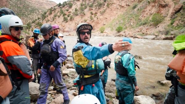A group of river rescuers in safety gear stand on rocky riverbank, with one person gesturing toward the water as they prepare for a rescue drill.