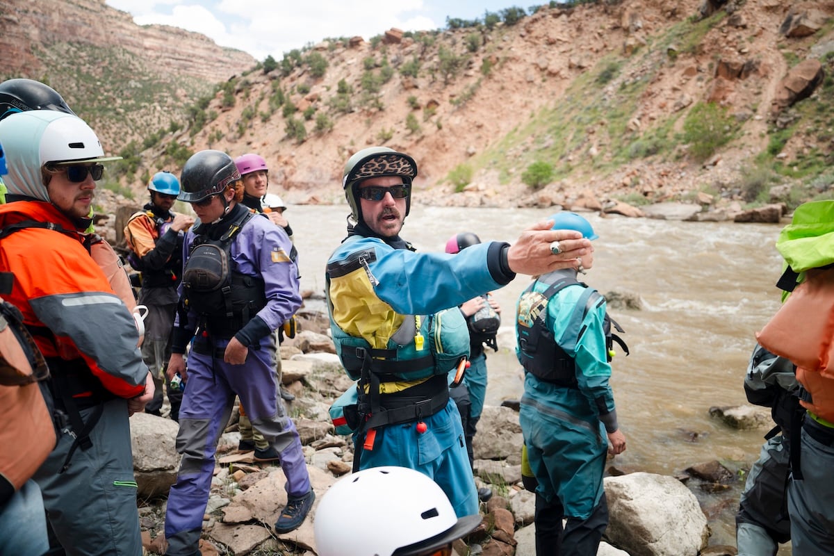 A group of river rescuers in safety gear stand on rocky riverbank, with one person gesturing toward the water as they prepare for a rescue drill.