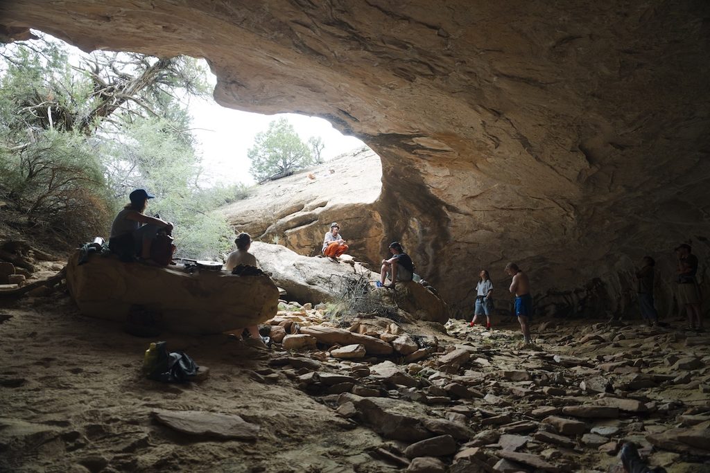 A group of people rests inside a large, rocky cave with natural light filtering through an opening above, surrounded by rugged terrain and scattered stones.