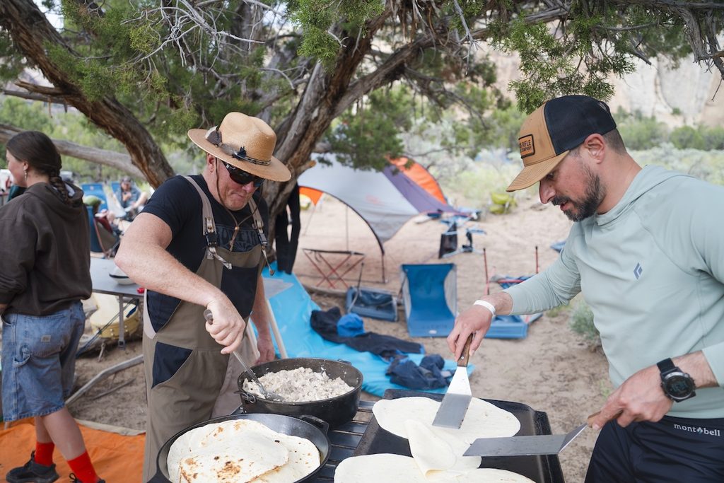 Two individuals prepare food outdoors under a tree, one stirring a pot and the other flipping tortillas on a griddle.