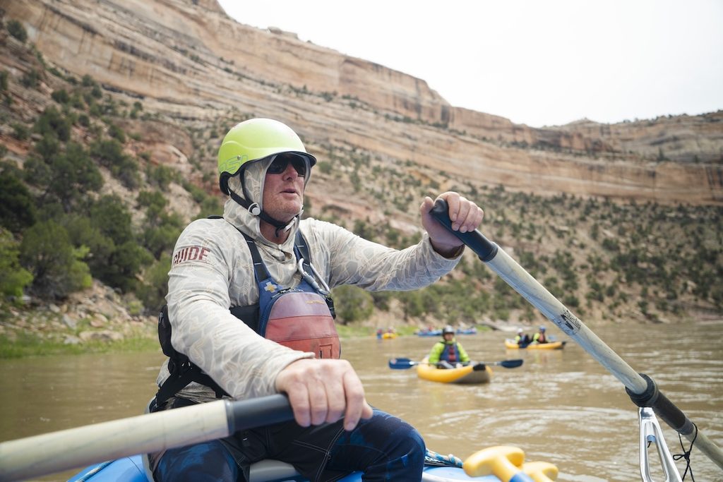 A person rows a raft a river, surrounded by towering rock cliffs and rafts and inflatable kayaks in the distance.