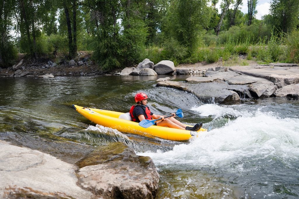 A person in a red helmet and life jacket paddles a bright yellow kayak through whitewater rapids in a river surrounded by trees and rocks.
