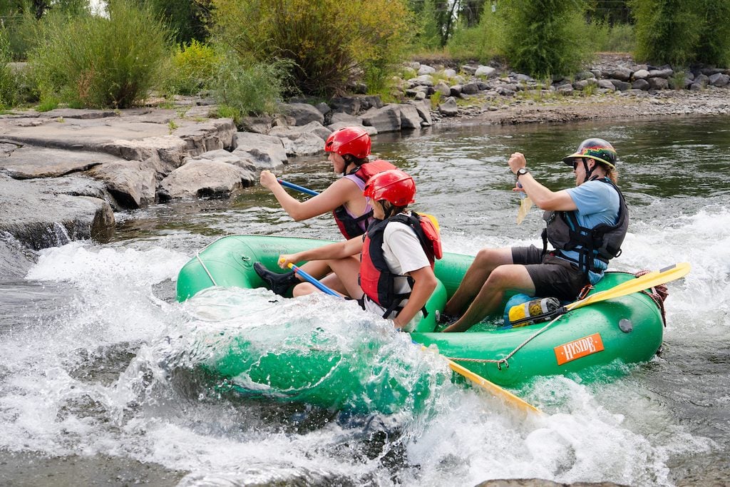 Three people in a green inflatable raft navigate whitewater rapids, paddling through turbulent waves with helmets and life vests.