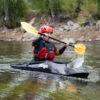 A person in a black kayak with a red helmet and life vest paddles through a river, splashing water as they navigate the current.