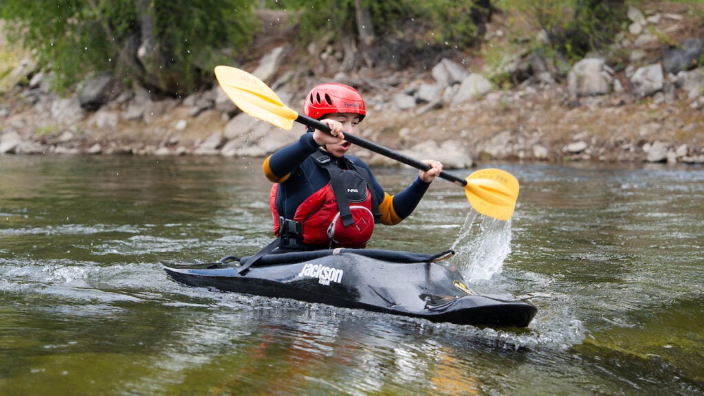 A person in a black kayak with a red helmet and life vest paddles through a river, splashing water as they navigate the current.
