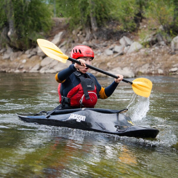 A person in a black kayak with a red helmet and life vest paddles through a river, splashing water as they navigate the current.