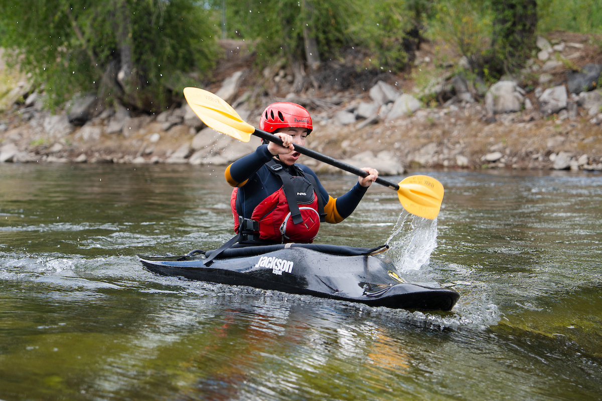 A person in a black kayak with a red helmet and life vest paddles through a river, splashing water as they navigate the current.